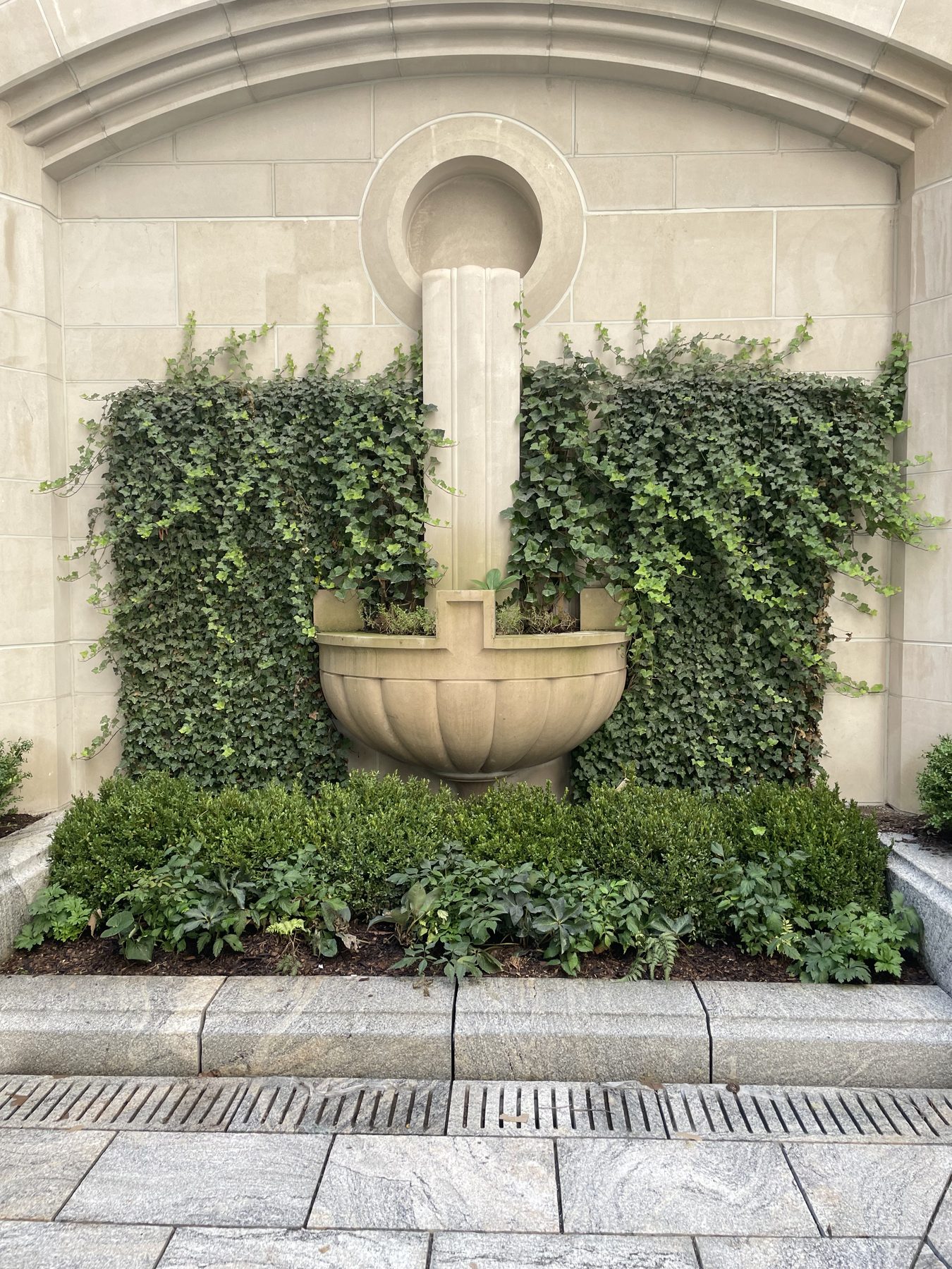A classical limestone wall fountain with English ivy growing in formal panels on either side, above a clipped boxwood and shade perennial bed.