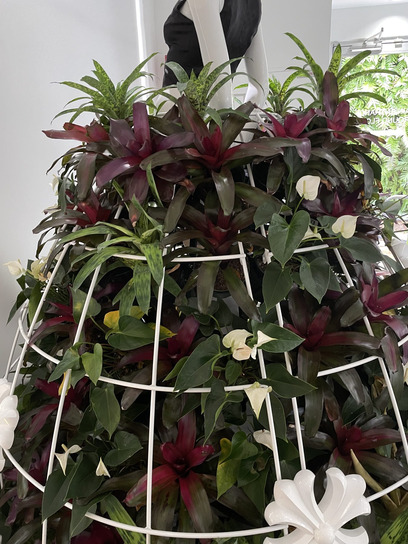 A floor-to-ceiling sculptural planter of bromeliads and anthurium installed on a white wire frame inside a retail store.